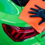 Ręka w czarnej rękawiczce z pomarańczową mikrofibrą poleruje zielony lakier samochodu. To część procesu dekontaminacji, co to jest?