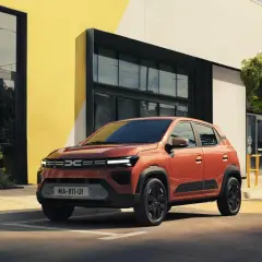Pomarańczowy Dacia Spring stoi przed nowoczesnym budynkiem. Sprawdź atrakcyjną dacia spring cena!