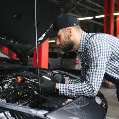 Mechanik w czapce i kratowanej koszuli bada silnik samochodu. Wygląda na to, że próbuje rozwiązać problem, który mógł wpędzić auto w **tryb awaryjny**.