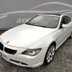 Białe BMW E63 coupe z jasnym wnętrzem, zaparkowane na żwirze, z logo autokomisu w tle.