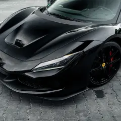 Czarny Ferrari F8 z błyszczącym lakierem i czerwonymi zaciskami hamulcowymi na czarnych felgach, zaparkowany na kostce brukowej.