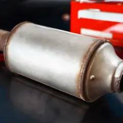Metalowy tłumik, element układu wydechowego, leży na ciemnej powierzchni. W tle widać fragment czerwonej skrzynki narzędziowej. To część do samochodu, być może do tuningu, jak nowy gpf.