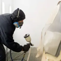 Lakiernik w kombinezonie i masce maluje samochód pistoletem lakierniczym.