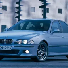 Błękitne e39 bmw z felgami M, zaparkowane przed nowoczesnym budynkiem.