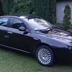 Czarna Alfa Romeo 159 lśni na trawie, otoczona zielenią drzew.