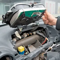 Dłoń majstruje przy desce rozdzielczej, przygotowując wymianę żarówki. Widoczne przewody i elementy mechanizmu.