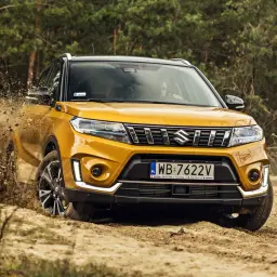 Żółty Suzuki Vitara Hybrid z impetem wjeżdża w błoto, tworząc kaskadę ziemi.