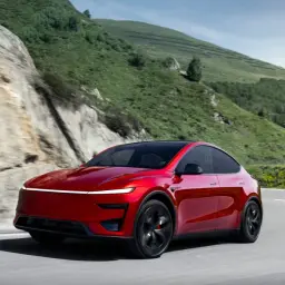 Czerwony Tesla Y Performance pędzi po krętej górskiej drodze, otoczony zielonymi zboczami i skalistymi klifami.