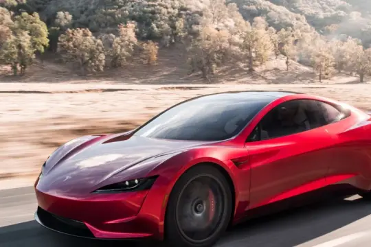 Czerwony Tesla Roadster pędzi po pustynnej drodze, otoczony suchymi drzewami i wzgórzami.