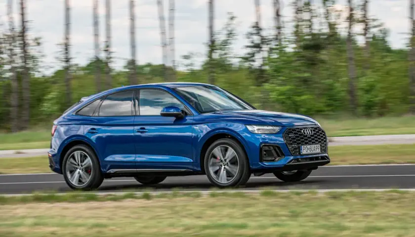 Błękitny Audi Q5 Sportback 2025 pędzi po drodze, otoczony zielenią i drzewami.