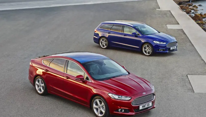 Dwa samochody Ford MK4: czerwony sedan i niebieskie kombi, stoją na asfaltowej nawierzchni przy betonowej konstrukcji i morzu.