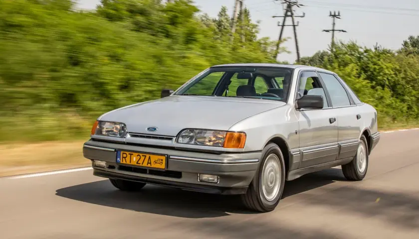 Srebrny Ford Scorpio jedzie po drodze, otoczony zielenią.
