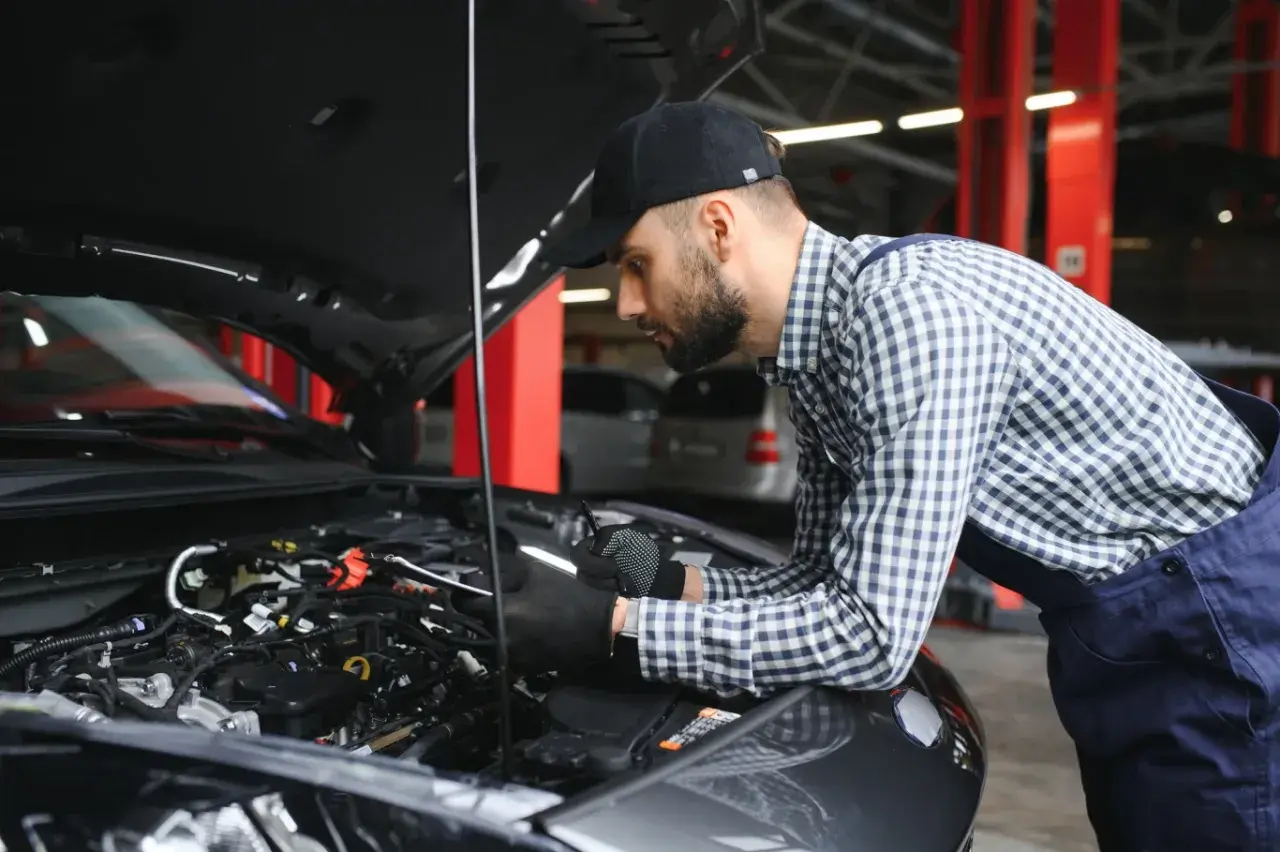 Mechanik w czapce i kratowanej koszuli bada silnik samochodu. Wygląda na to, że próbuje rozwiązać problem, który mógł wpędzić auto w **tryb awaryjny**.