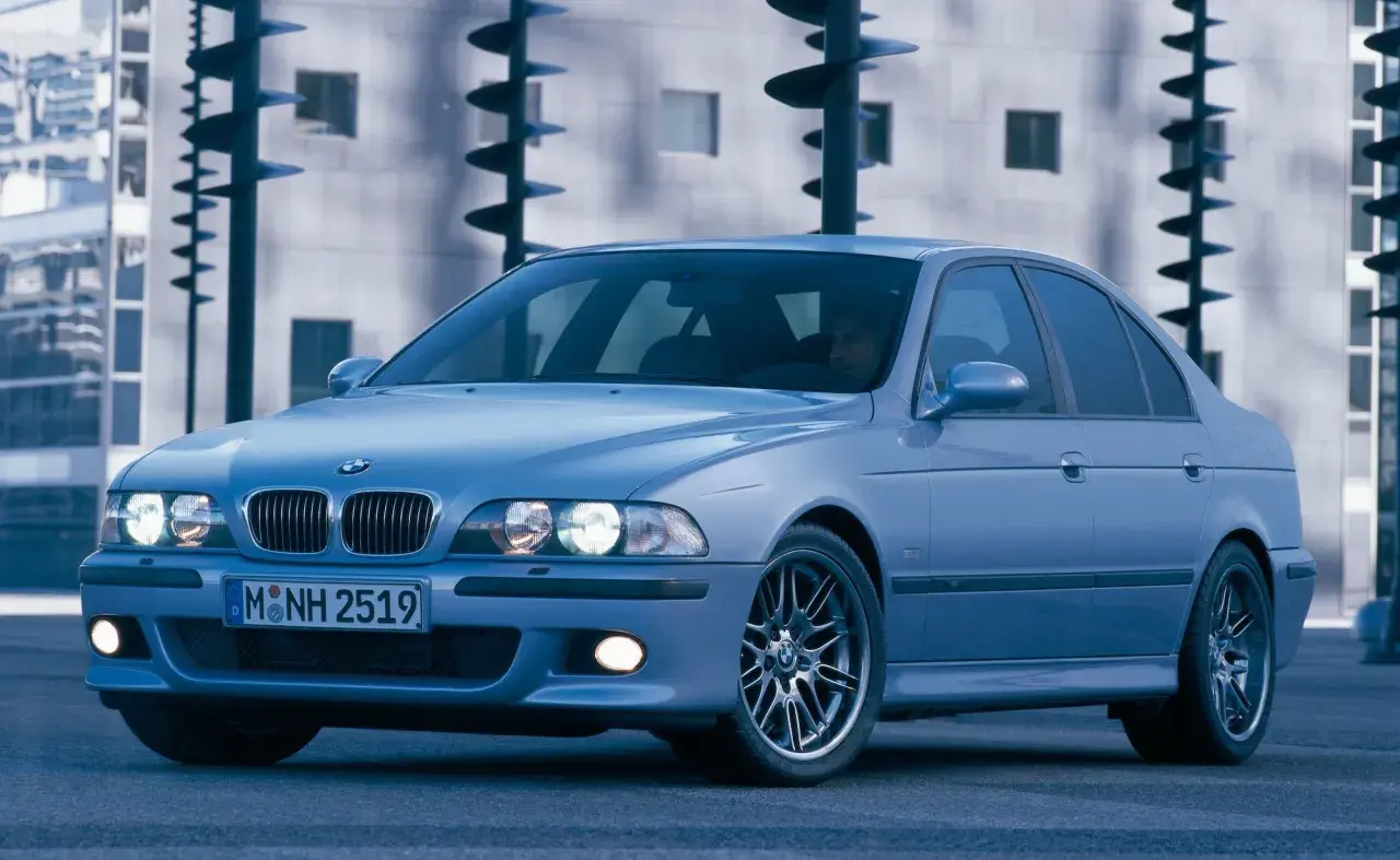 Błękitne e39 bmw z felgami M, zaparkowane przed nowoczesnym budynkiem.