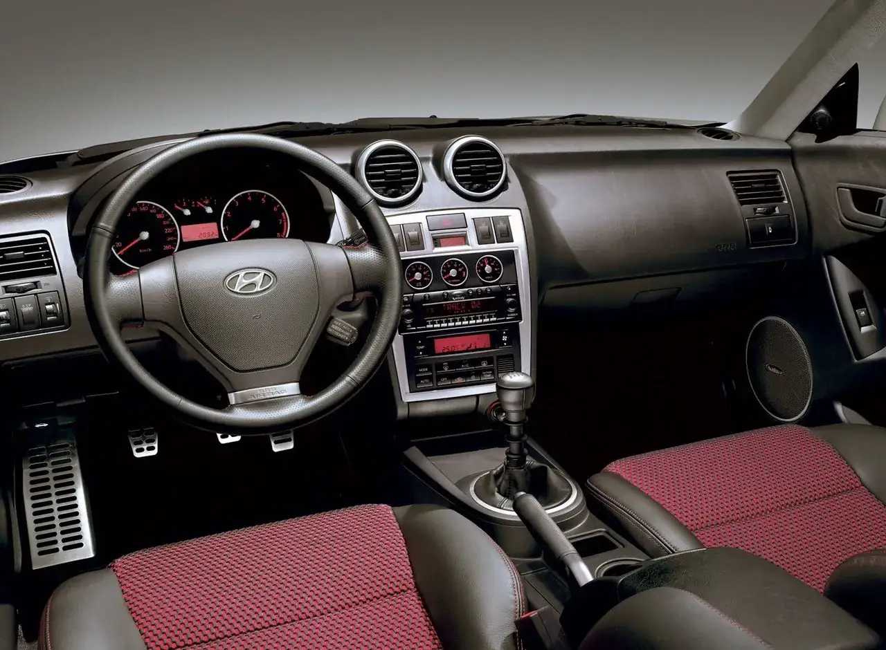 Wnętrze Hyundai Coupe 2.7 V6: kierownica, deska rozdzielcza z radiem i nawiewami, dźwignia zmiany biegów, sportowe fotele z czerwonymi wstawkami.